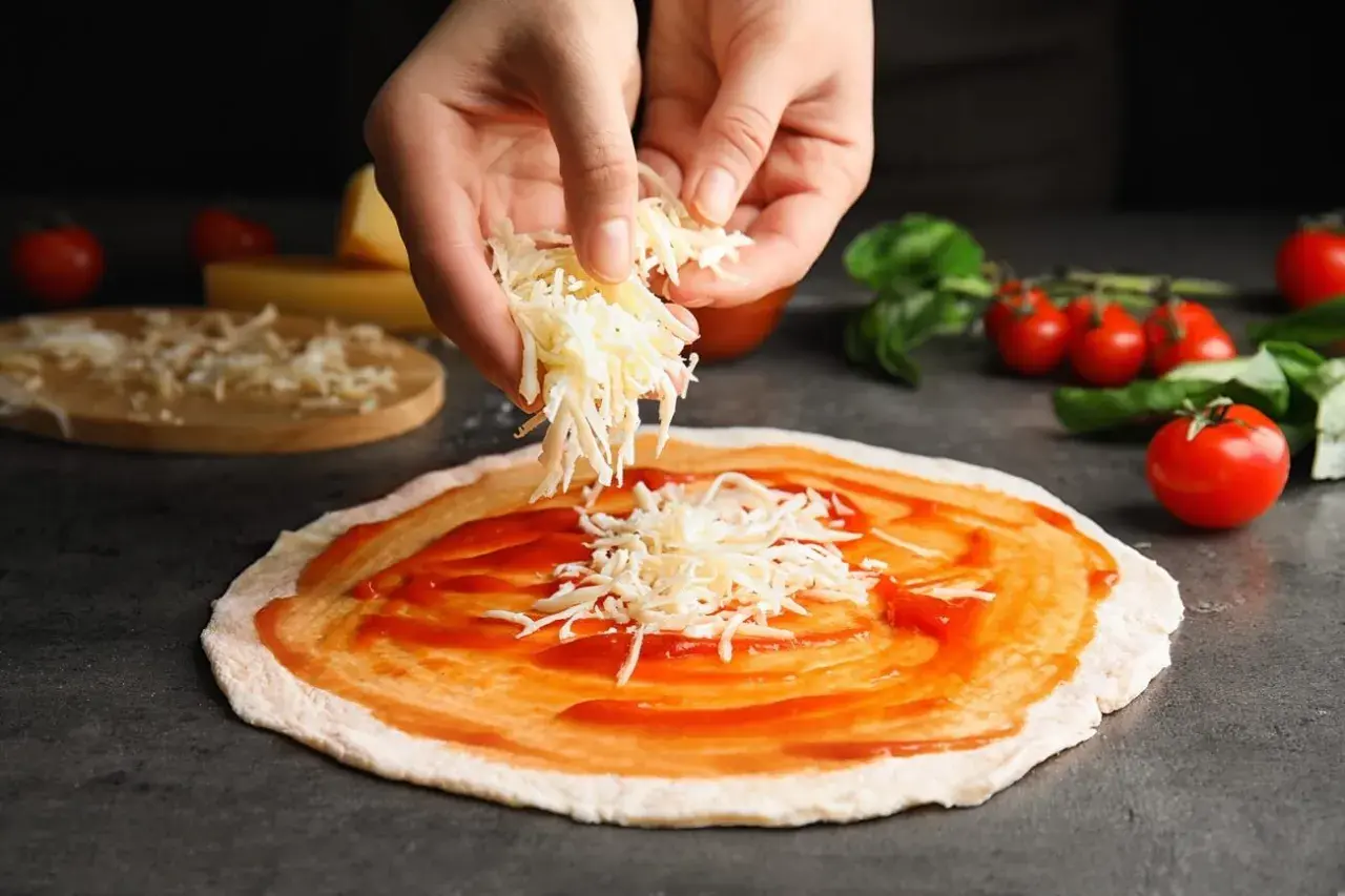 Hände streuen geriebenen Gouda auf eine mit Tomatensauce bestrichene Pizza. Tomaten und Basilikum liegen daneben.