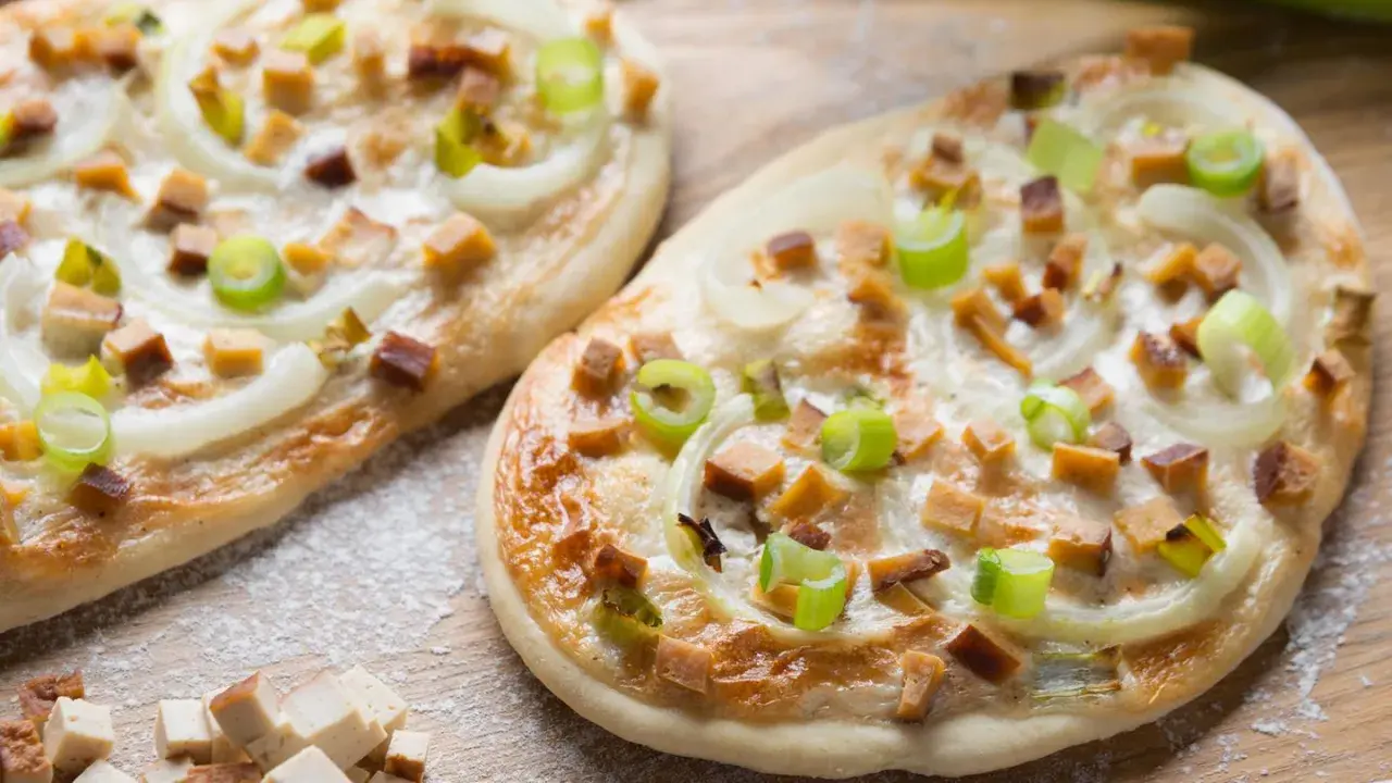 Zwei knusprige, vegetarische Flammkuchen mit Zwiebeln, Frühlingszwiebeln und würzigen Tofuwürfeln, bestreut mit etwas Mehl.