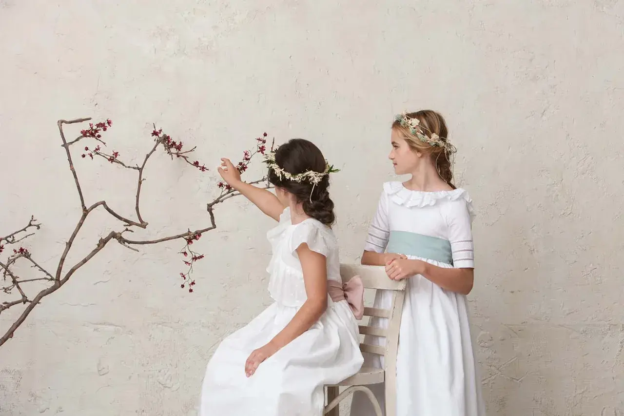 Dos niñas con peinados de comunión con diadema floral, una sentada tocando ramas, la otra de pie.