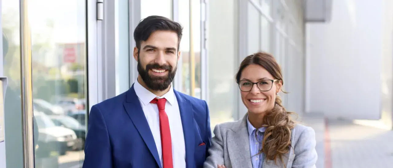 Smiling professionals, a man in a blue suit and red tie, and a woman in a grey blazer, stand outside. They're ready to pursue online MPA programs no GRE.