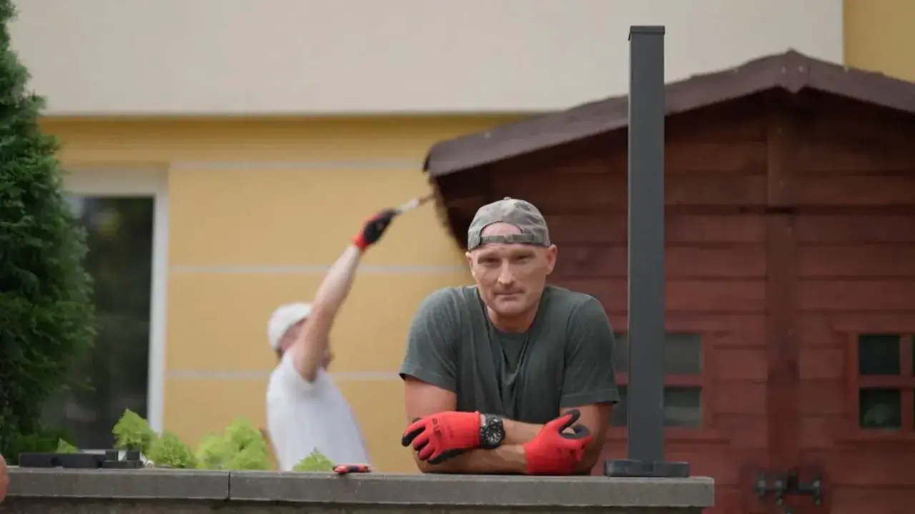 Mężczyzna w czapce i rękawiczkach opiera się o mur. W tle inny mężczyzna maluje drewnianą szopę. Jak zgłosić się do polowania na ogród?
