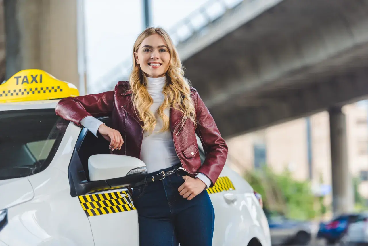 Uśmiechnięta blondynka w skórzanej kurtce opiera się o białe taxi. Chcesz wiedzieć, jak zostać kierowcą Opti Taxi?