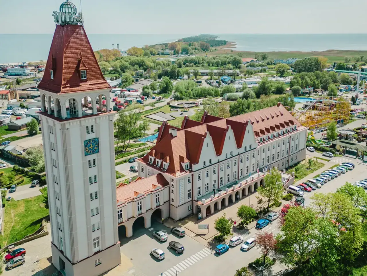 Wieża zegarowa i długi budynek z czerwonym dachem, przypominający dom rybaka, stoją obok siebie. W tle widać morze i park rozrywki.