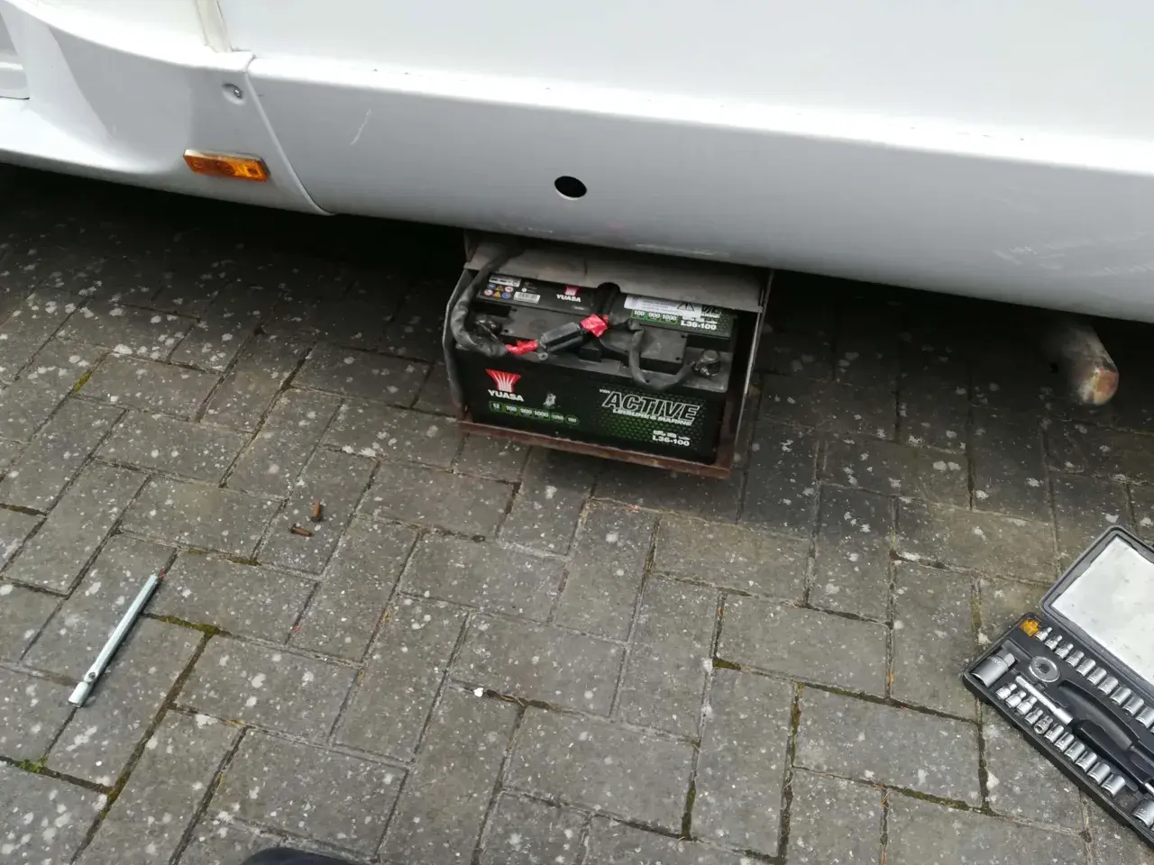 Emplacement batterie Ford Transit : une batterie Yuasa "Active Leisure & Marine" est visible sous le véhicule, dans un support métallique. Une clé à douille et une boîte à outils sont au sol.
