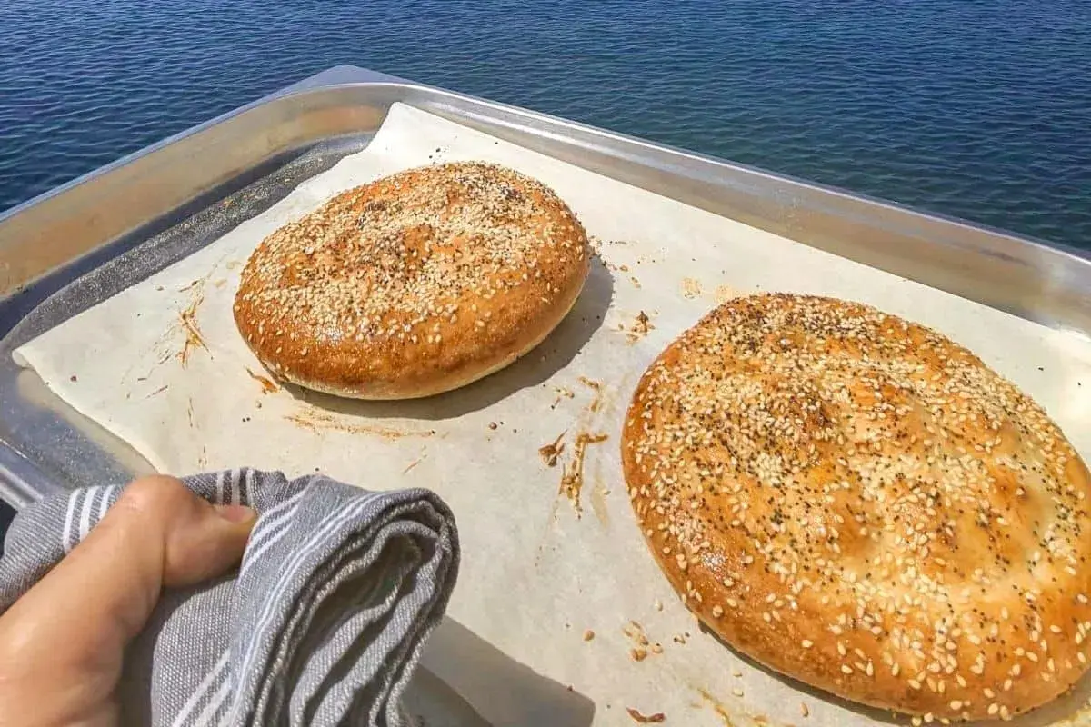 Två gyllene ramazan pidesi, toppade med sesamfrön, vilar på ett bakplåtspapper vid havet.