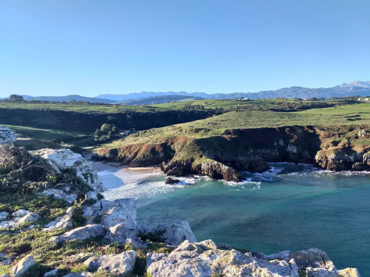 Acantilados y mar turquesa, ideales para rutas senderismo San Vicente de la Barquera. Paisaje costero con verdes colinas y montañas al fondo.