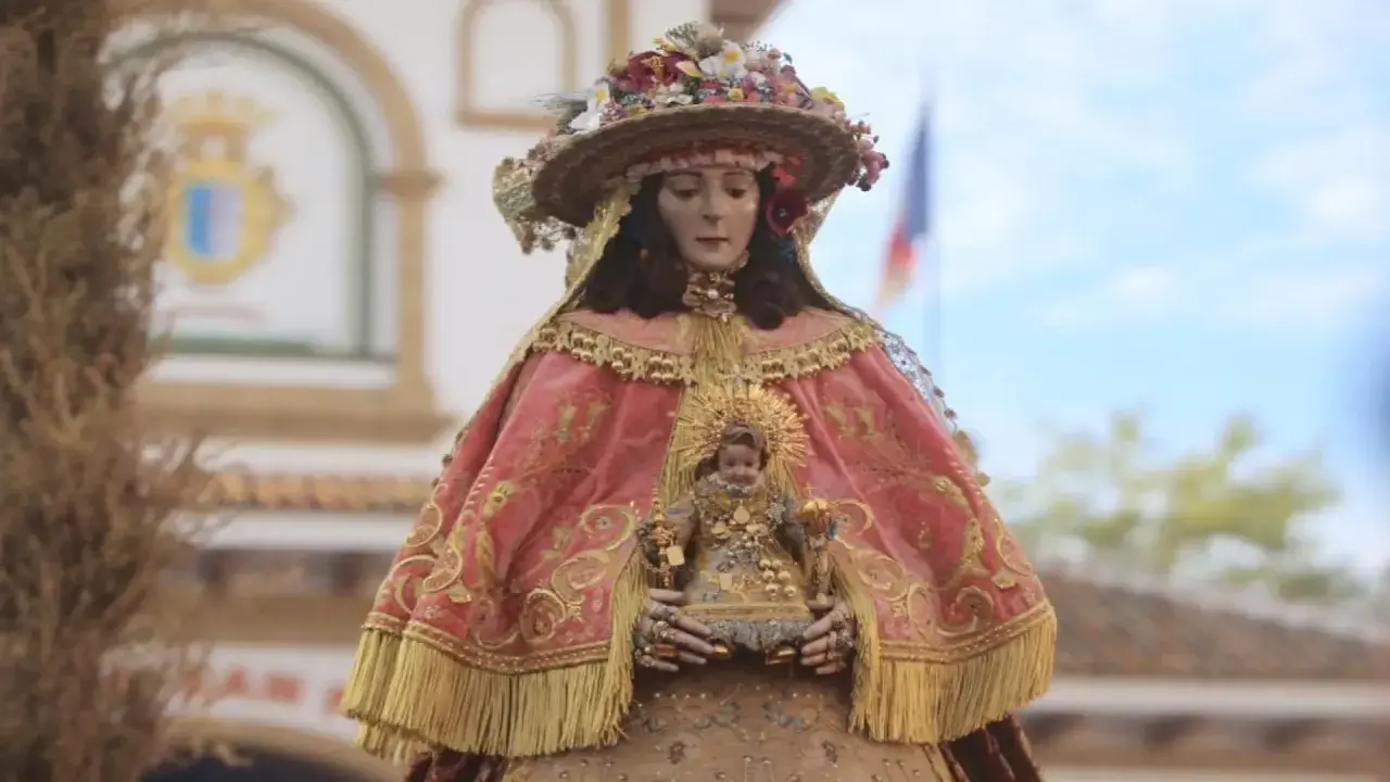 Virgen del Rocío pastora, con su manto rosa bordado y el Niño Jesús en brazos, adornada con flores y un sombrero.
