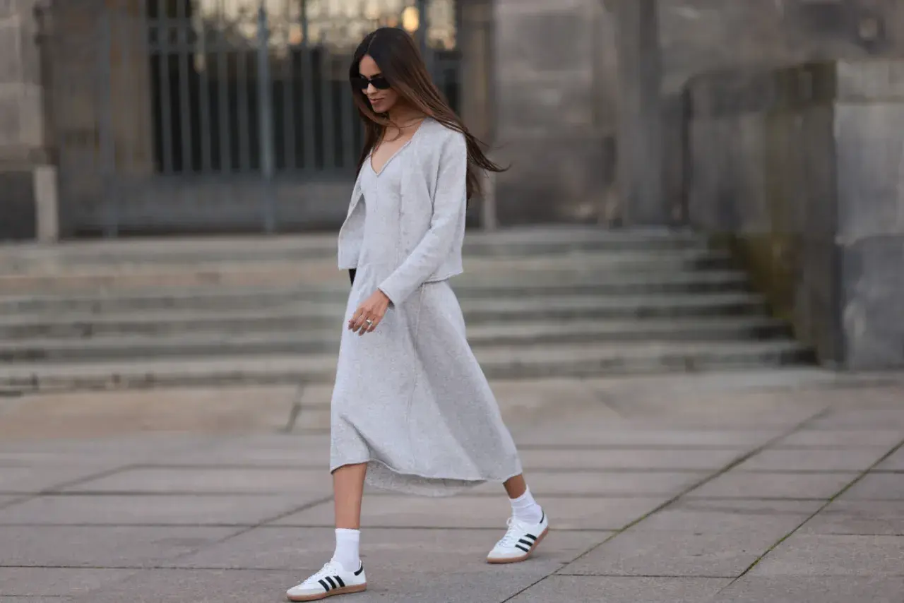 Elegancka sukienka i sportowe buty tworzą stylowy kontrast. Kobieta w szarym komplecie i białych sneakersach idzie ulicą.