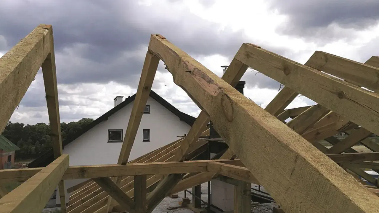 Montaż więźby dachowej w toku. Drewniane belki tworzą konstrukcję dachu nad białym domem z kominem.