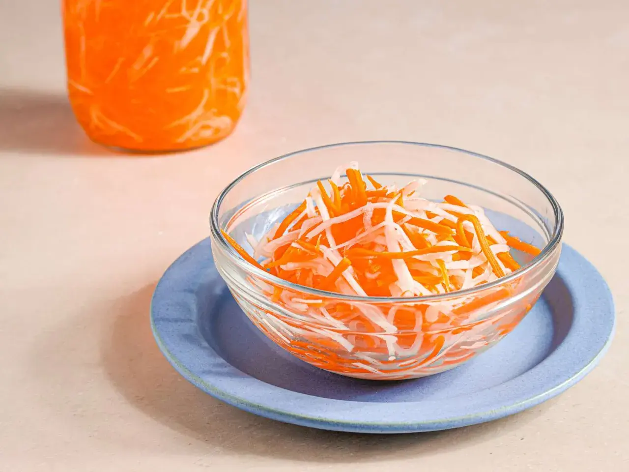 A bowl of vibrant japanese pickled carrots and daikon radish sits on a blue plate, with a jar of the same pickled vegetables in the background.