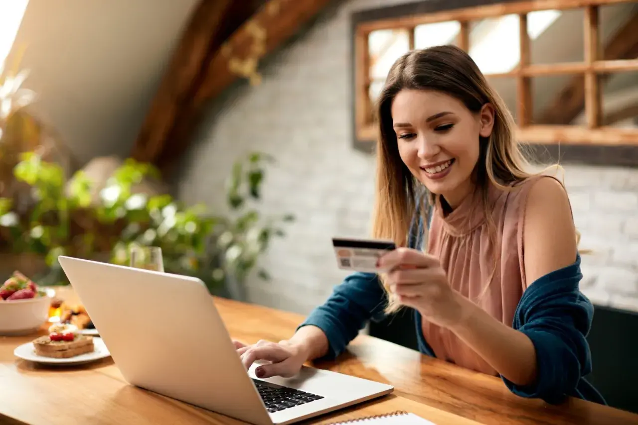 Uśmiechnięta kobieta z kartą płatniczą przy laptopie, planuje zakupy. Sprawdza cenę netto przed finalizacją transakcji.