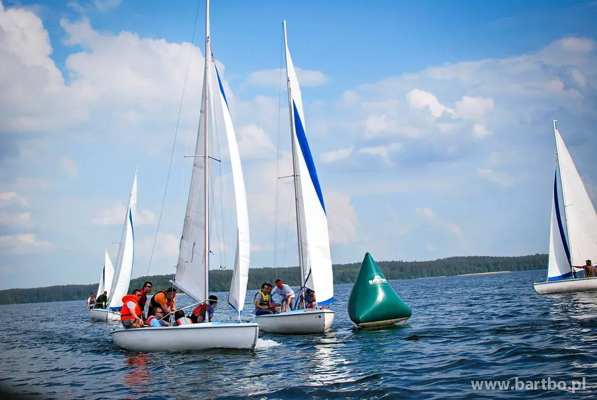 Regata żeglarska na jeziorze. Kilka łodzi z załogami ściga się, mijając zieloną bojję.