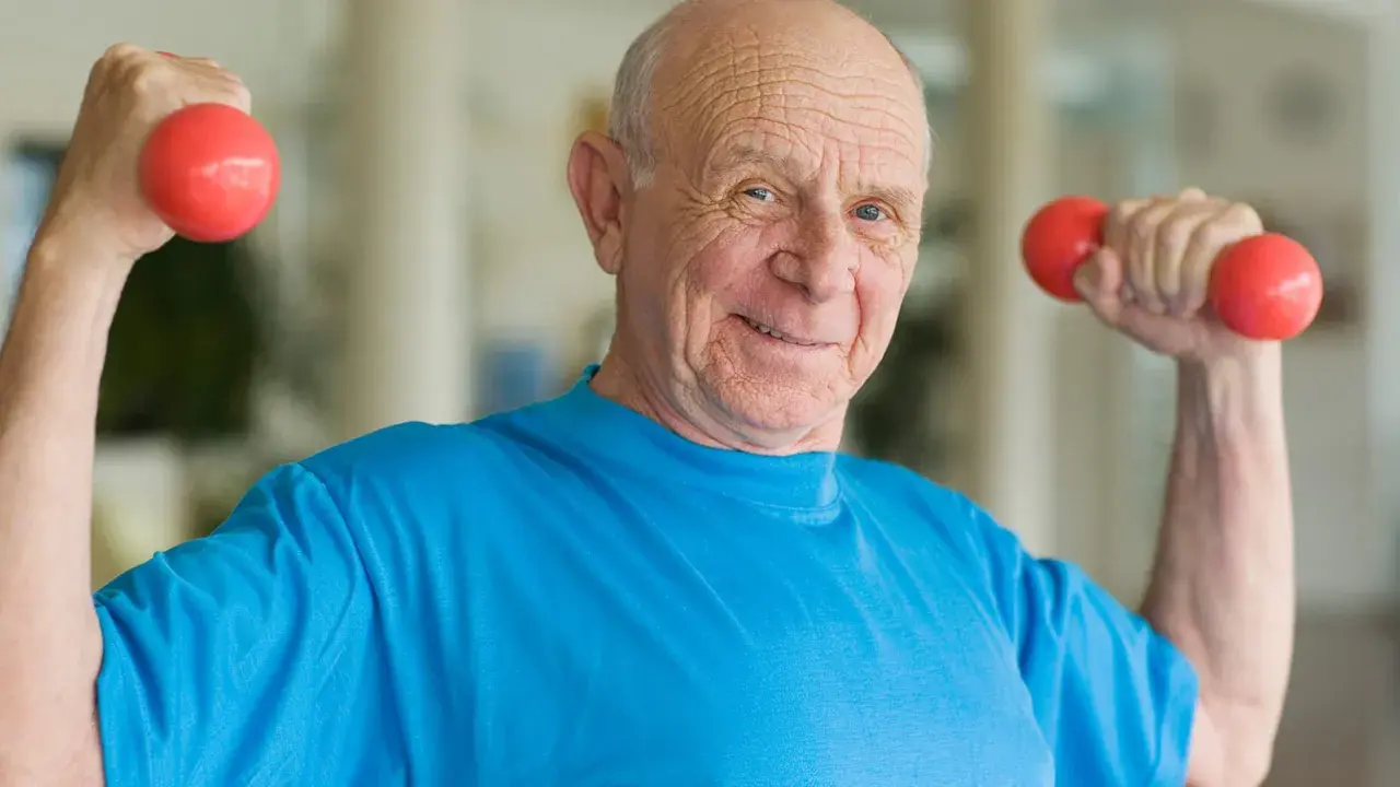 Ein lächelnder älterer Mann macht Muskeltraining im Alter mit roten Hanteln.