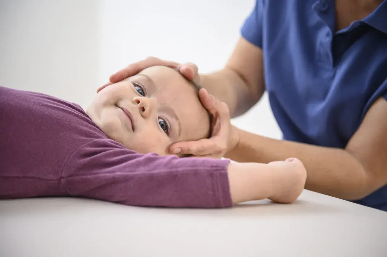 Sanfte Hände einer Osteopathin untersuchen den Kopf eines lächelnden Kleinkindes.