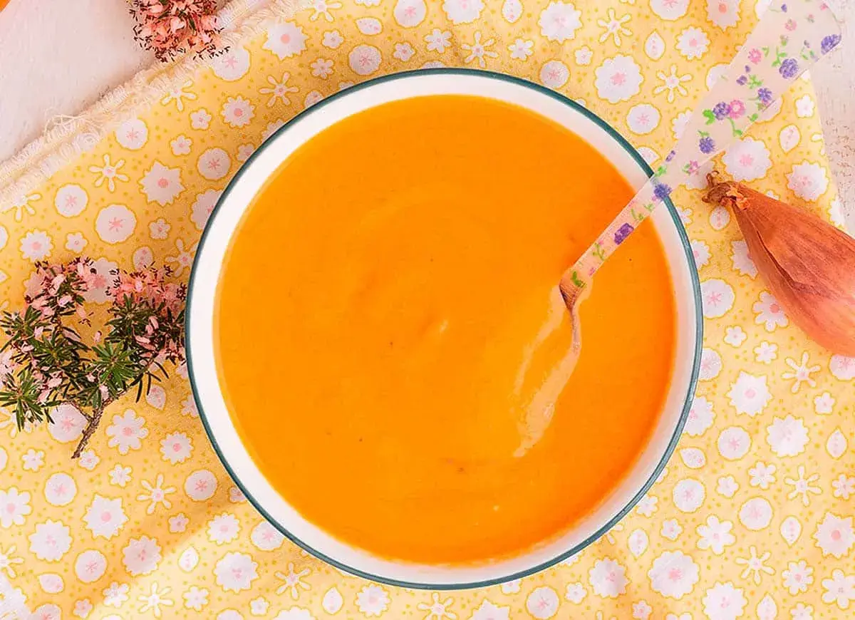 Un tazón de cremosa salsa americana, lista para disfrutar. Una cuchara con flores y una chalota completan la escena.