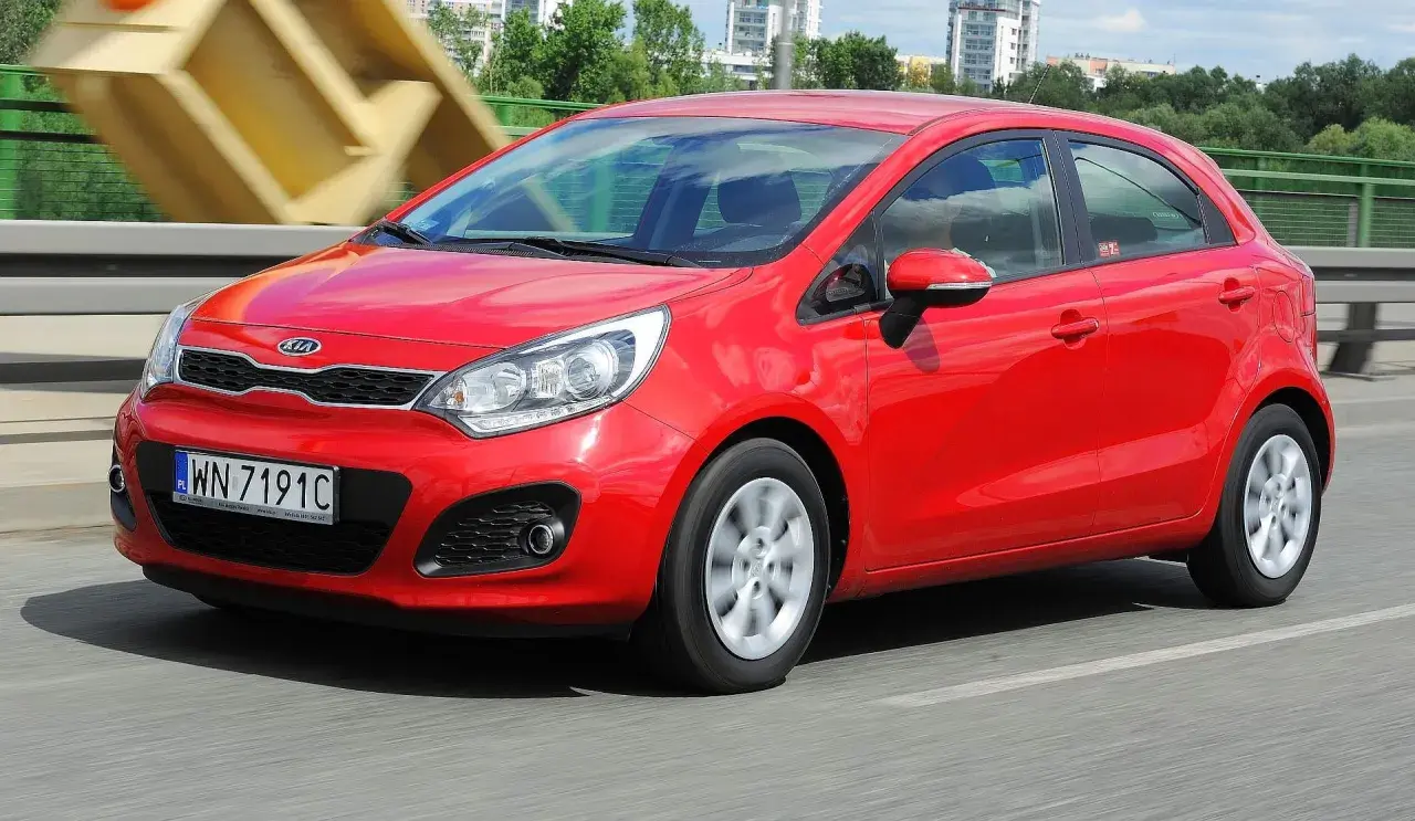 Czerwony Kia Rio 3 w ruchu, z widocznym numerem rejestracyjnym WN 7191C, na tle mostu i budynków.