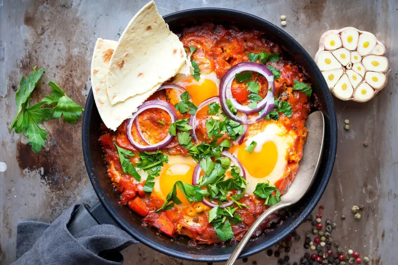 Délicieuse recette de chakchouka dans une poêle, garnie d'œufs, d'oignons rouges et de persil frais. Servi avec du pain pita.