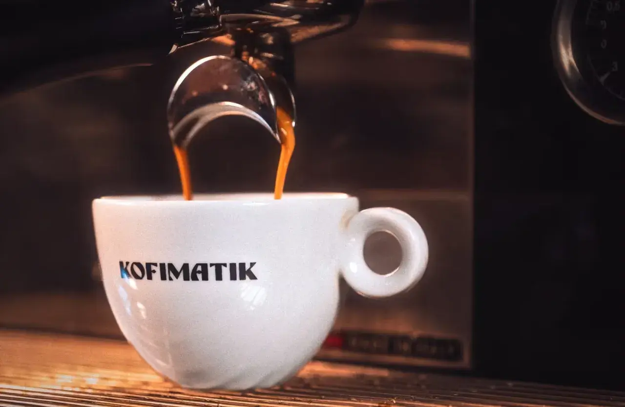 Gorąca ristretto kawa spływa do białej filiżanki z logo Kofimatik. Intensywny aromat i głęboki smak.