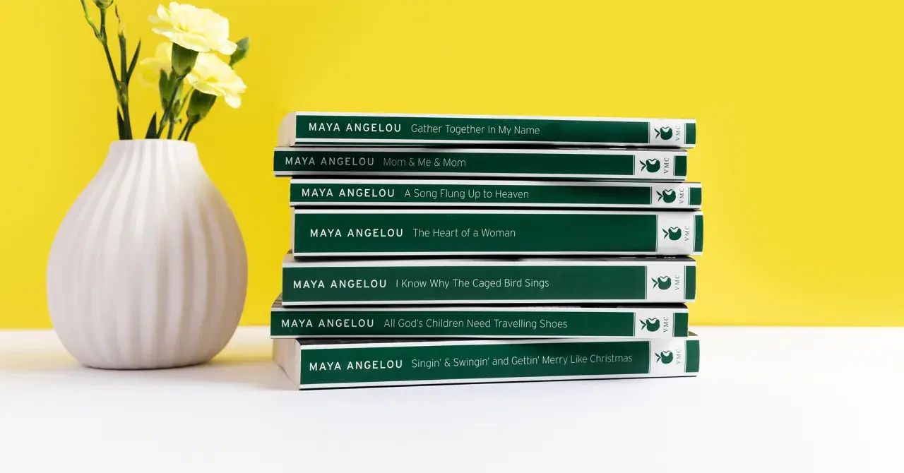 Stos **książek** **Maya Angelou** obok wazonu z żółtymi kwiatami.