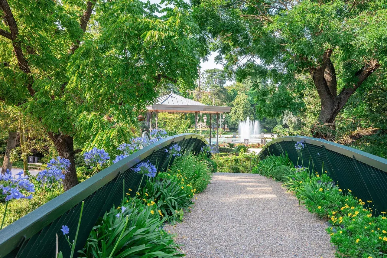Un pont fleuri mène à un kiosque et une fontaine au jardin des plantes de Toulouse.