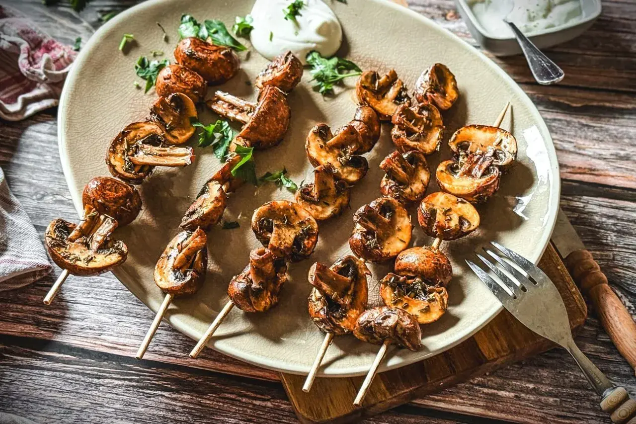 Gegrillte Champignons auf Spießen, garniert mit Kräutern und einem Klecks Dip. Perfekt für ein sommerliches BBQ.