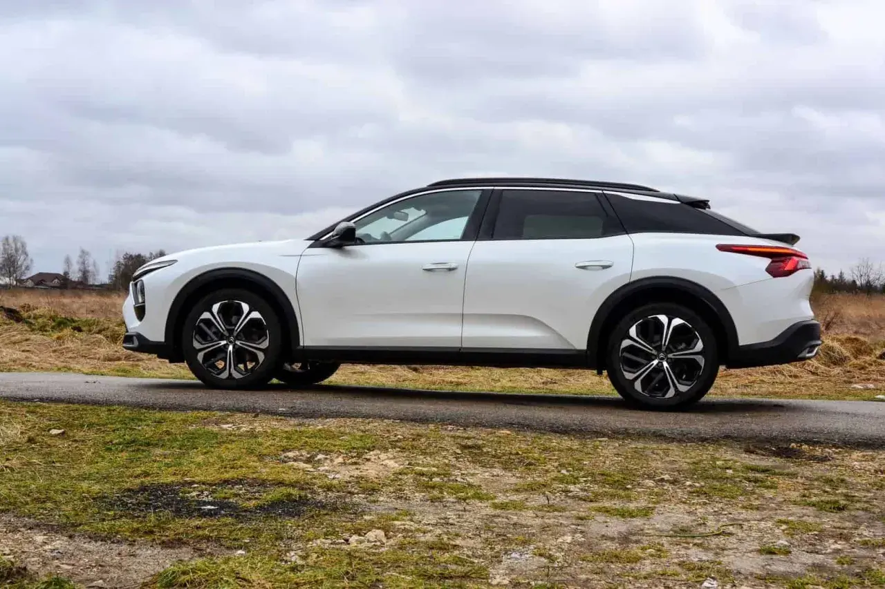 Biały Citroen C5X na tle pochmurnego nieba i wiejskiego krajobrazu. Elegancki design i nowoczesne felgi podkreślają jego wyjątkowość.