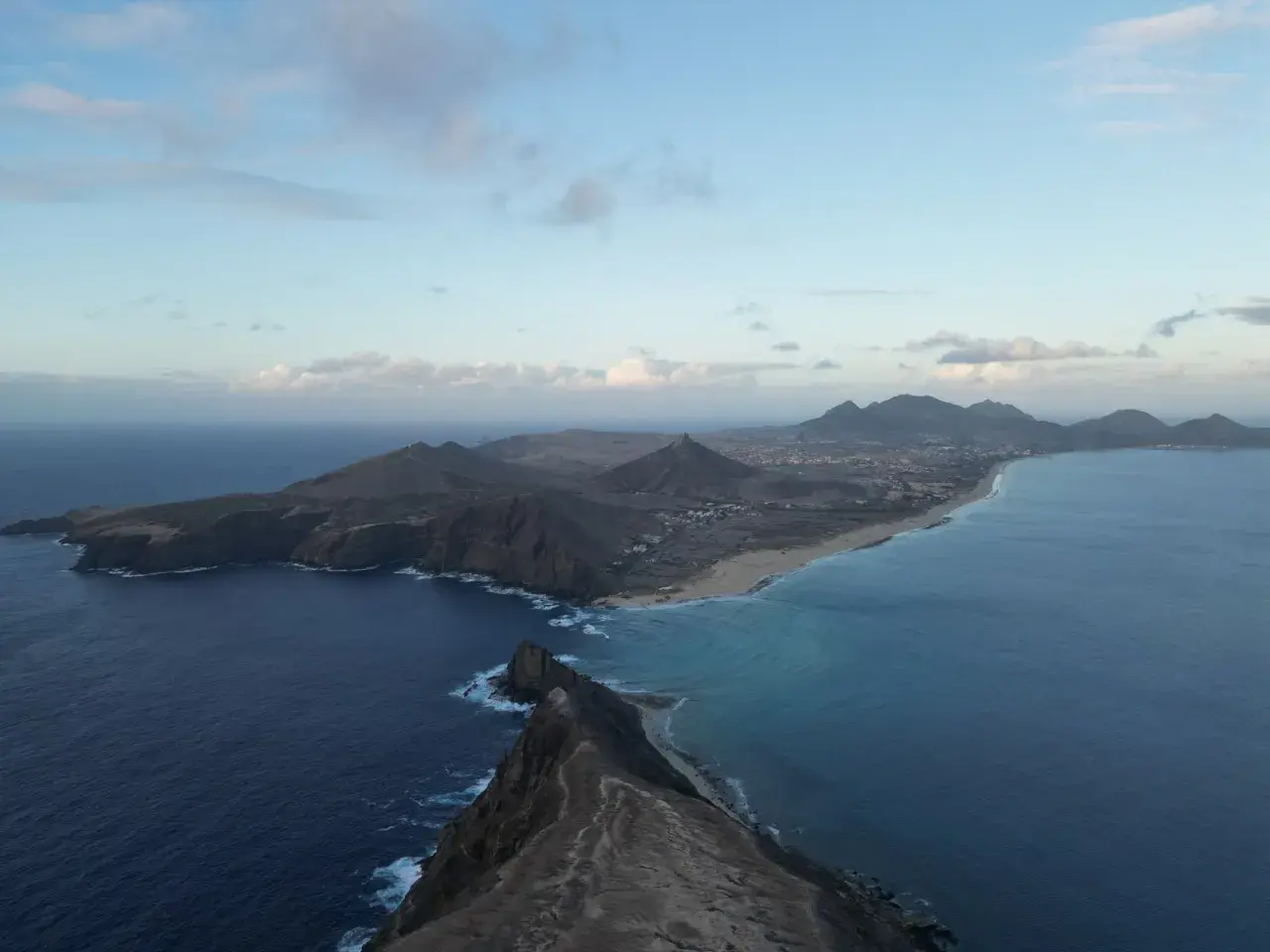 Dramatic coastline of Porto Santo island, with rugged cliffs meeting turquoise waters and a long sandy beach stretching towards distant mountains under a cloudy sky.