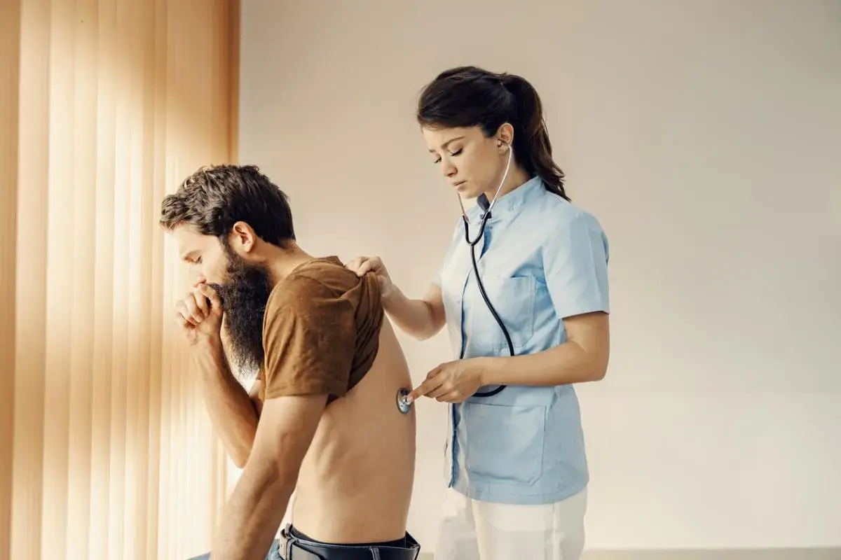 Médica examina a un hombre con barba que tose, sintiendo su espalda. Podría tener dolor de espalda al toser.
