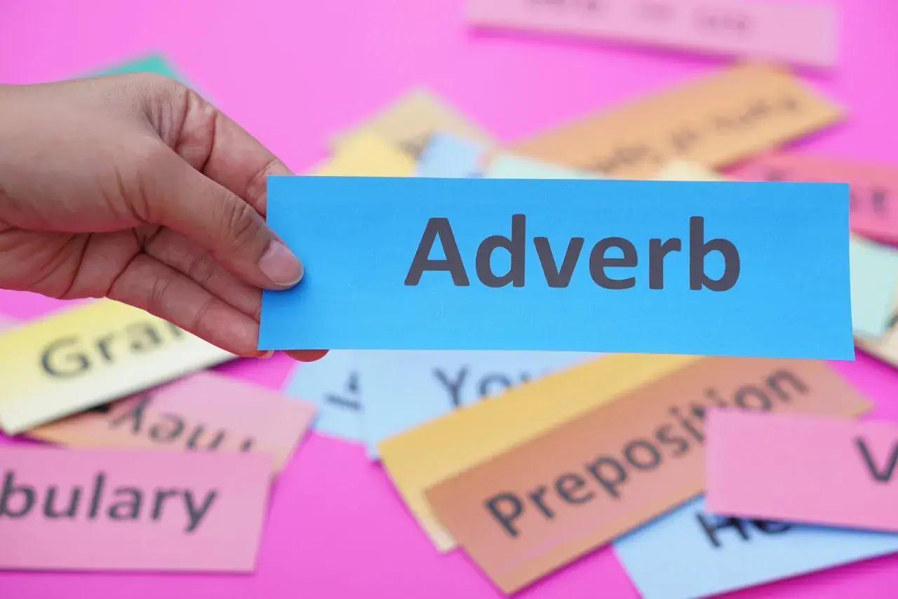 Una mano tiene un biglietto blu con la parola "Adverb". Sullo sfondo, altri biglietti colorati con parole come "Grammar" e "Vocabulary".
