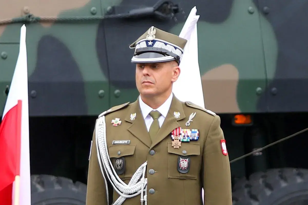 Oficer w mundurze, z orderami na piersi, stoi obok polskiej flagi. Nad nim widać fragment pojazdu wojskowego.