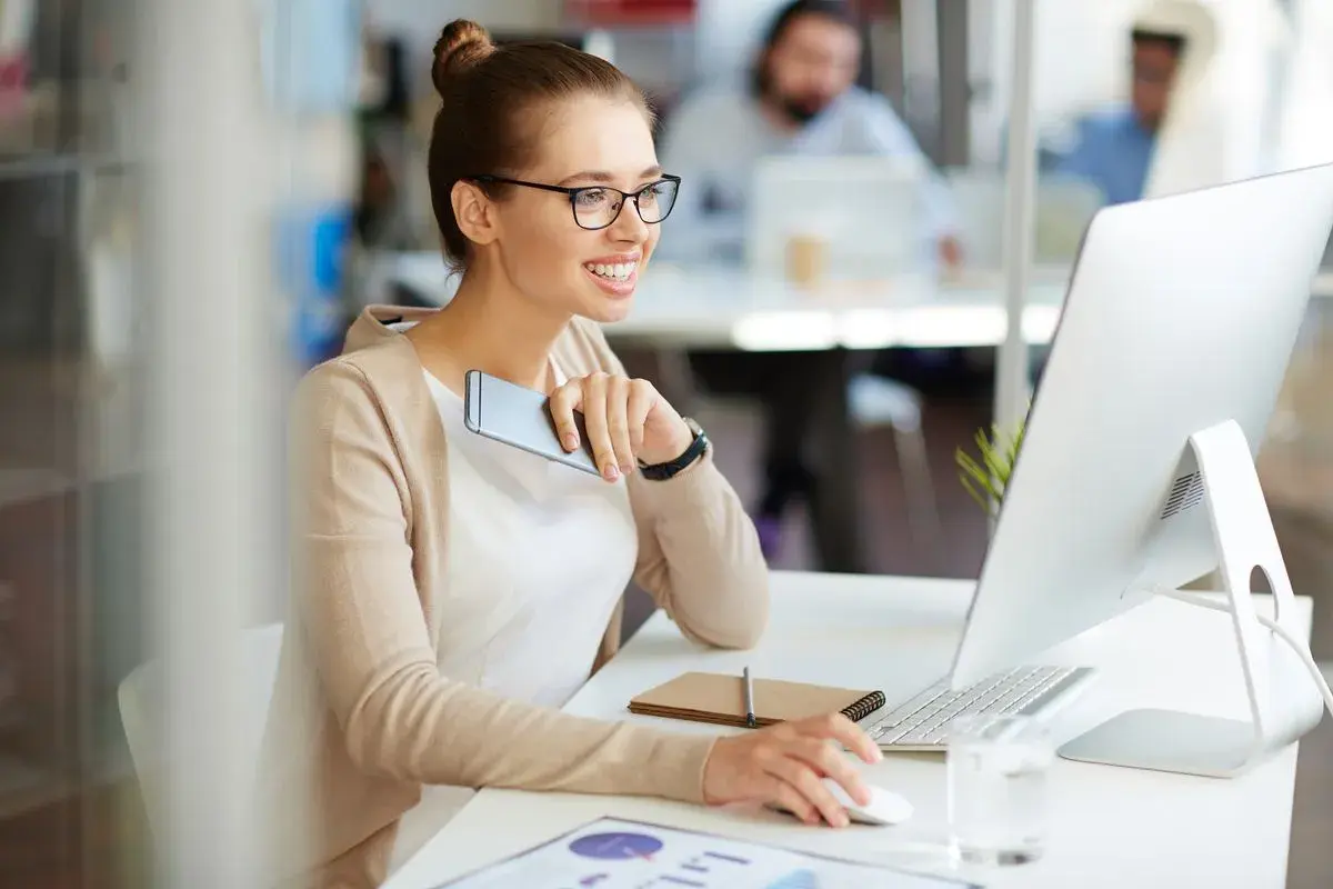 A smiling woman in glasses works at a computer, holding a phone.