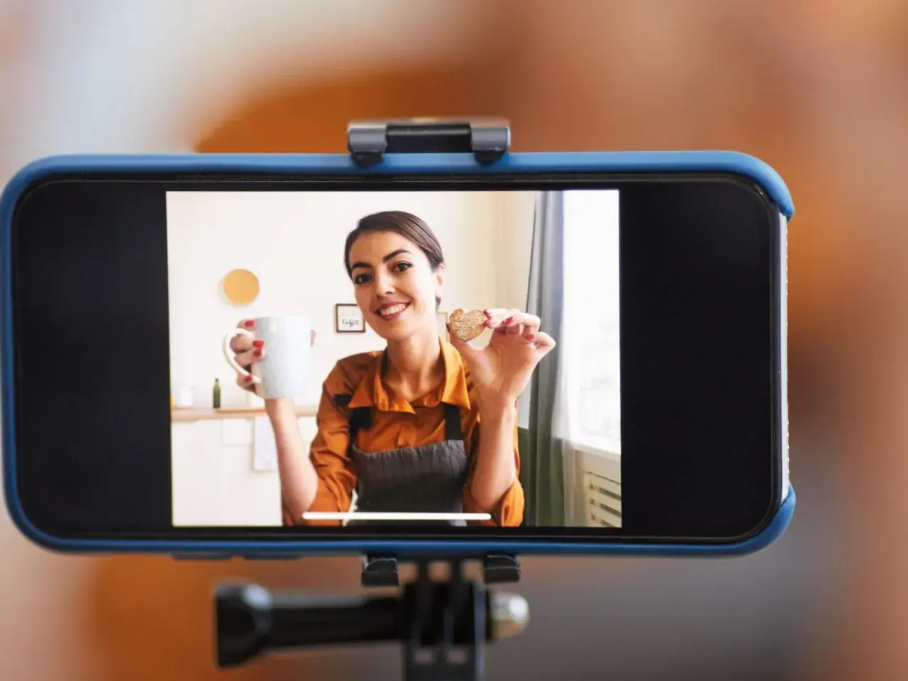 Frau hält Tasse und Keks, während sie ihr **iPhone als Webcam** für ein Kochvideo nutzt.