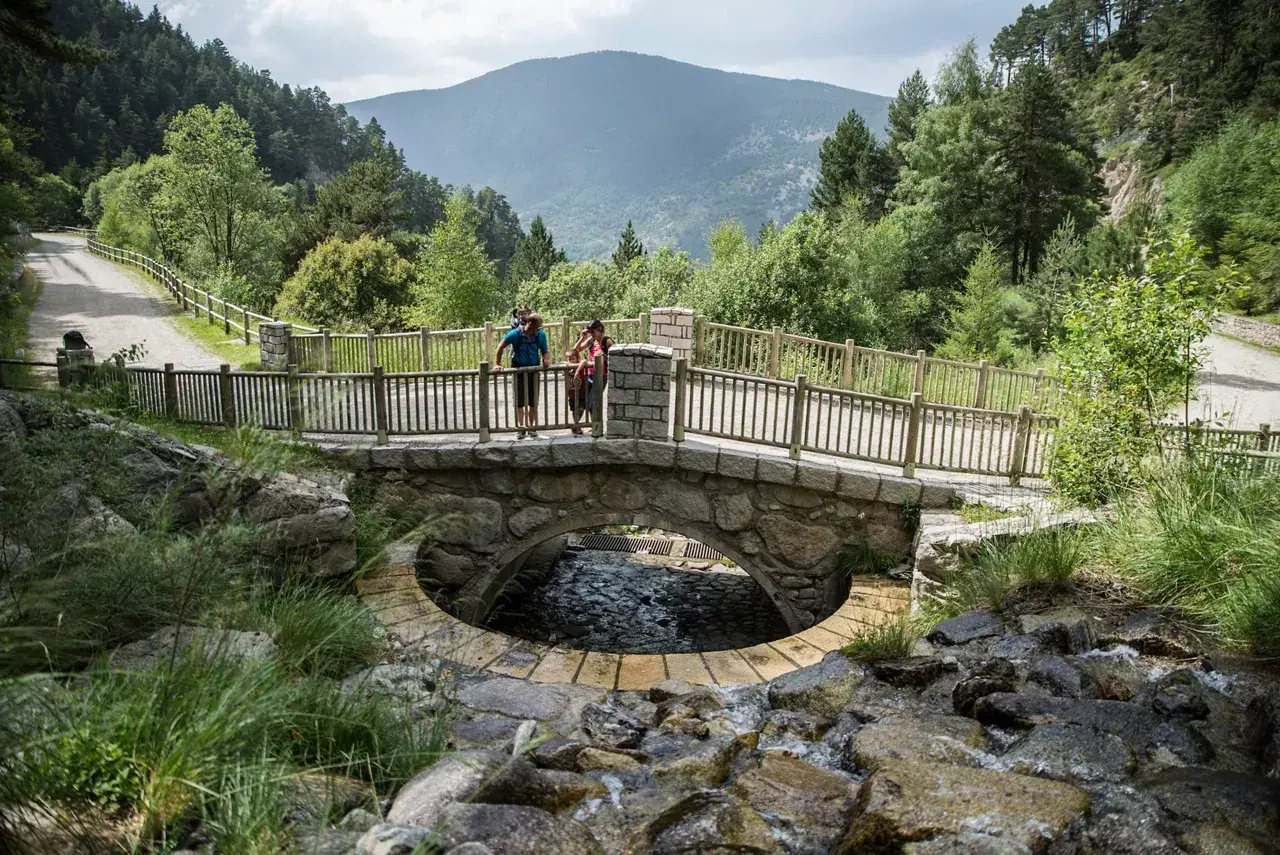 Pareja en un puente de piedra sobre un arroyo en Les Pardines, Andorra. Montañas y bosques al fondo.