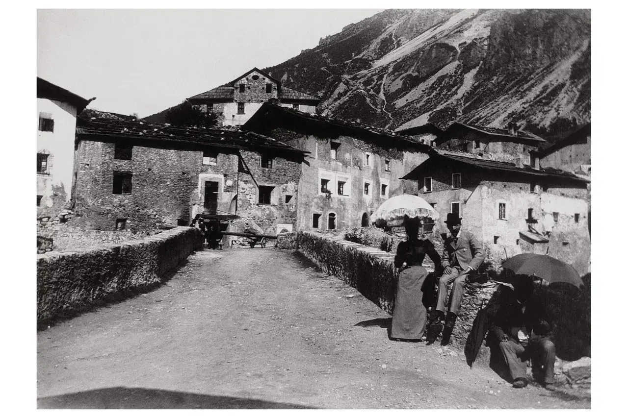 Un villaggio di montagna immortalato da Giovanni Verga fotografo. Coppie eleganti passeggiano con ombrelli, sullo sfondo case in pietra e pendii scoscesi.