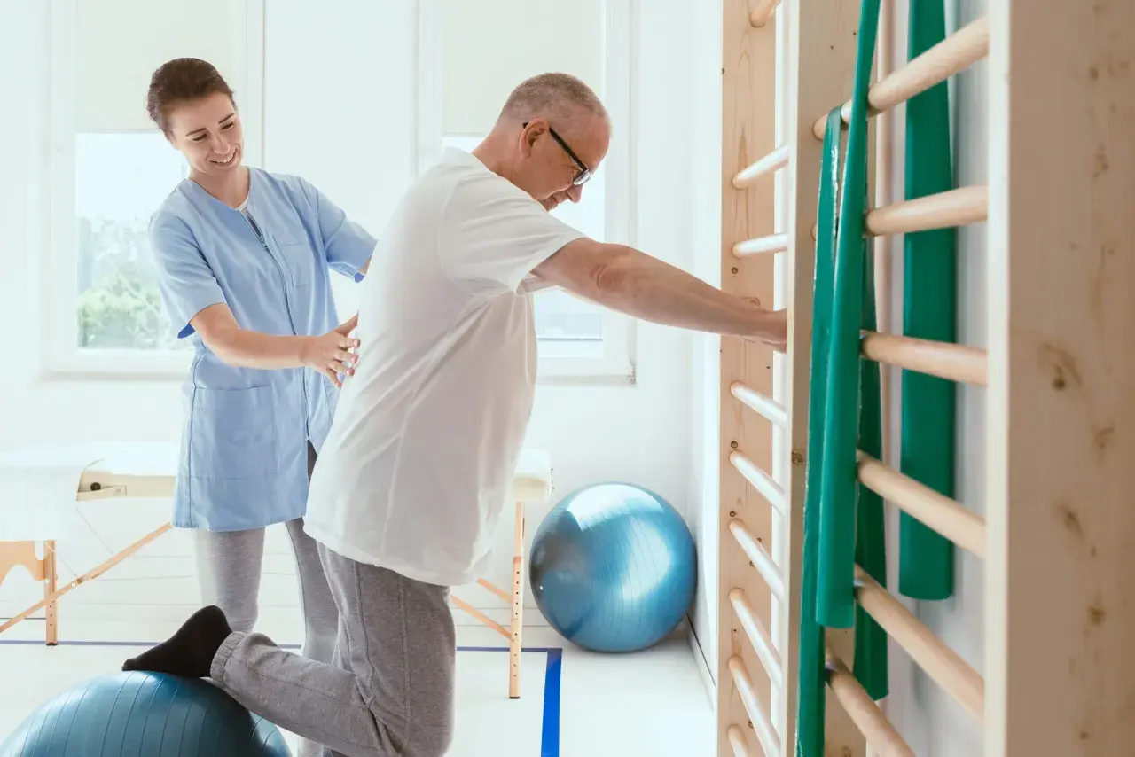 Fizjoterapeutka pomaga pacjentowi ćwiczyć na piłce rehabilitacyjnej przy drabinkach.