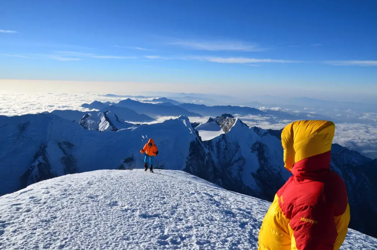 Widok z góry, gdzie są Himalaje. Dwóch alpinistów w kolorowych kurtkach na ośnieżonym szczycie, pod nimi morze chmur i kolejne pasma górskie.