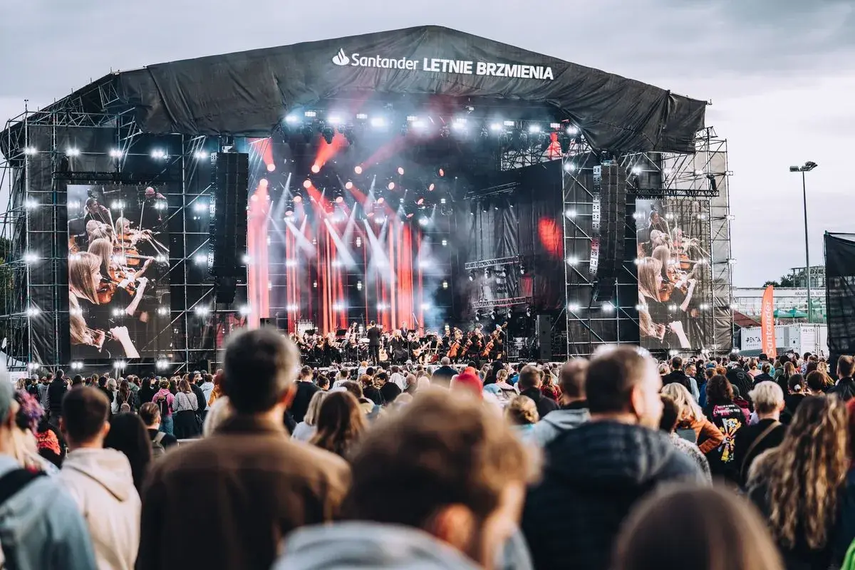 Koncert Santander Letnie Brzmienia z orkiestrą na żywo i ekranami pokazującymi muzyków. Tłum widzów czeka na występ.