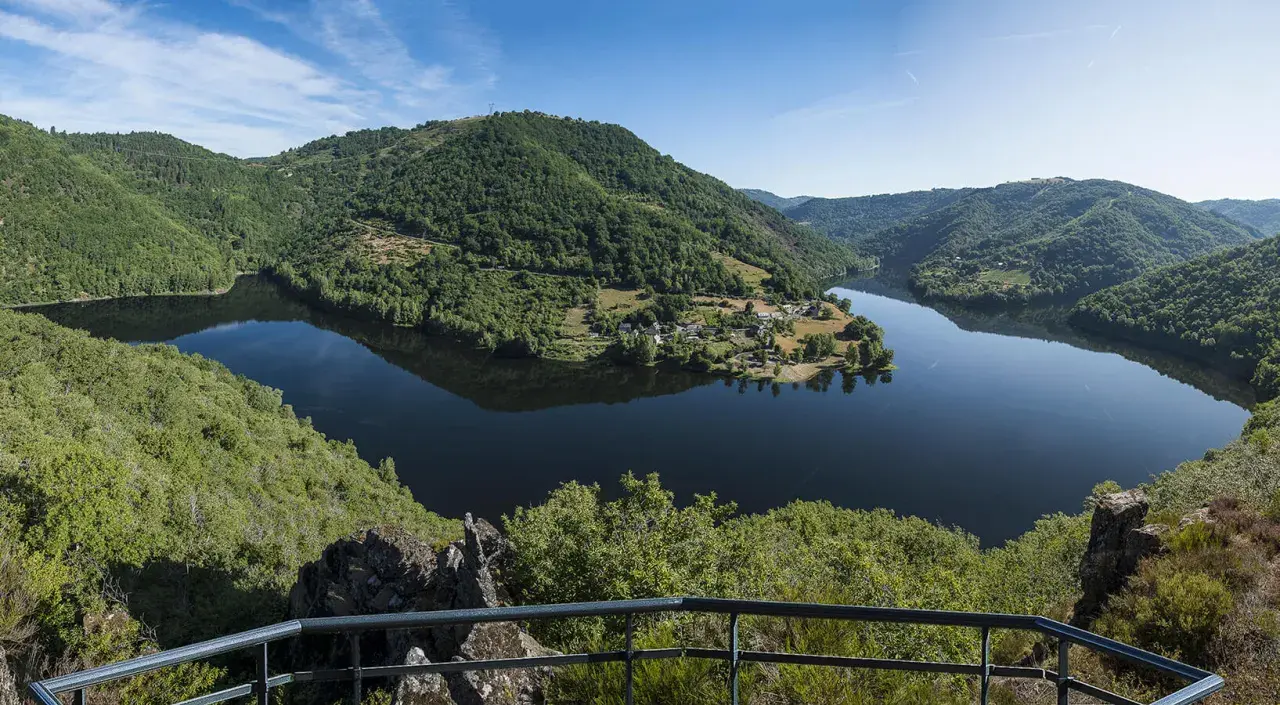 Vue panoramique des gorges de la Truyère, avec un lac paisible reflétant les collines verdoyantes et un village pittoresque.