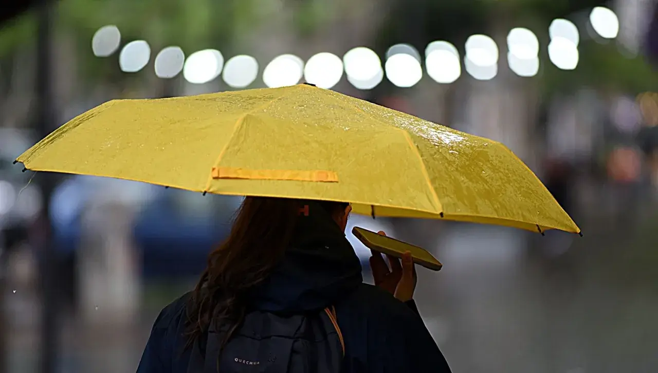 Una persona con un paraguas amarillo, bajo la lluvia, parece soñar con un día soleado mientras habla por teléfono.