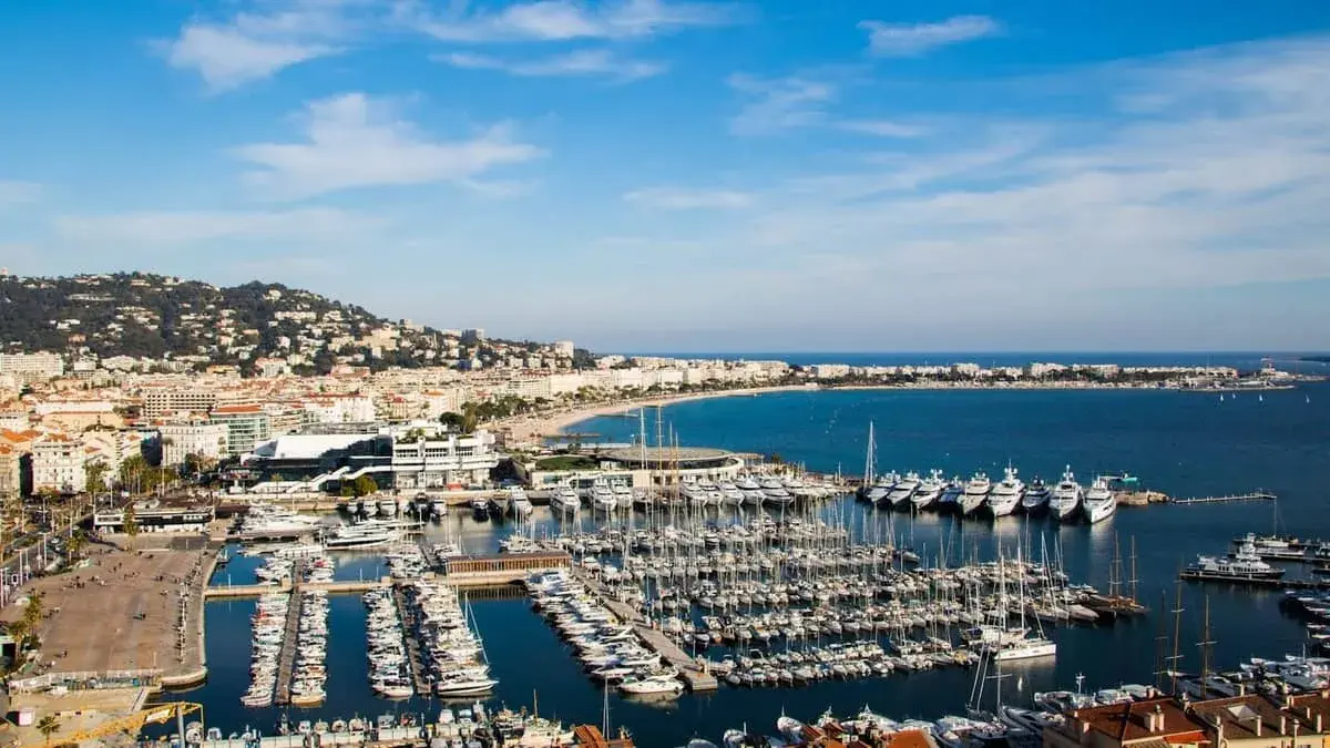 Widok na port w Cannes, gdzie cumują luksusowe jachty. W tle rozciąga się plaża i wzgórza. Idealne miejsce, by zobaczyć Cannes.