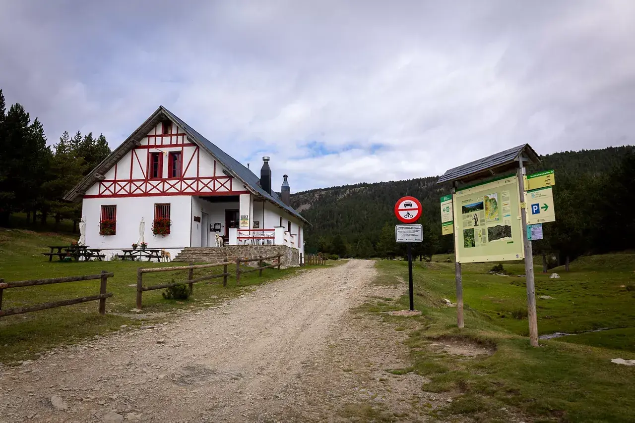 Refugi comes de rubio, un refugio de montaña blanco con detalles rojos, rodeado de pinos y un camino de grava.