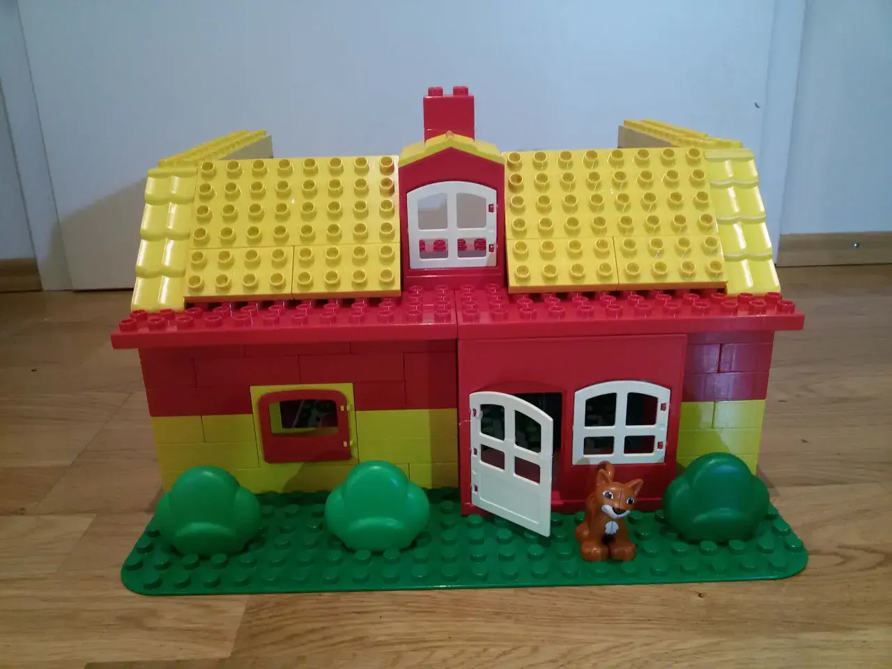 Ein farbenfrohes Lego Duplo Haus mit gelbem Dach, roten Wänden, Fenstern, einer Tür und einem kleinen Fuchs davor. Perfekt für kreative Bauanleitungen.