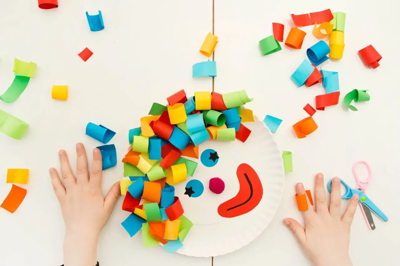 Bambini creano lavoretti di carnevale da appendere: un pagliaccio con capelli colorati su un piatto di carta, circondato da coriandoli di carta.
