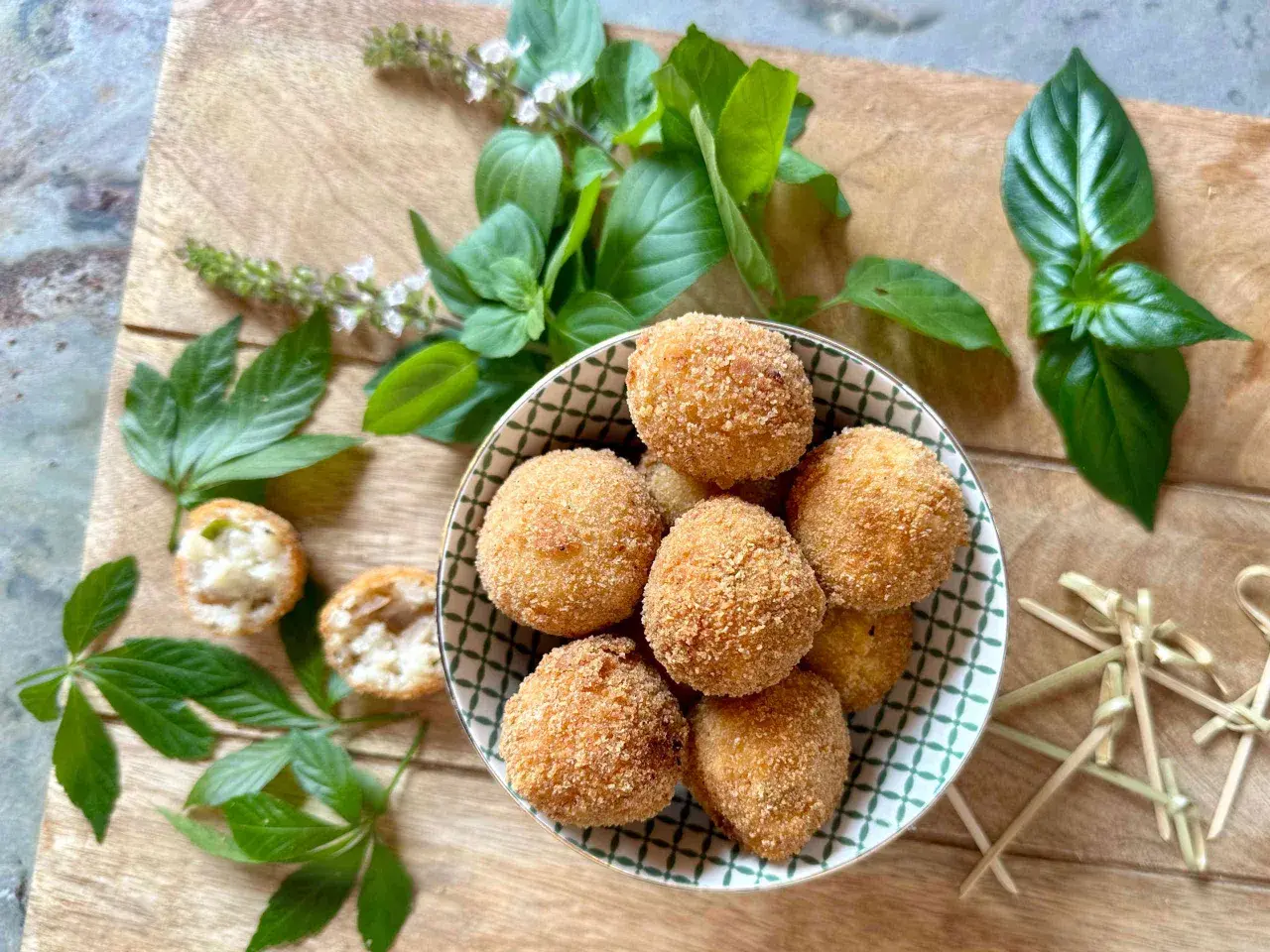 Goldbraune Arancini, frisch zubereitet nach einem tollen arancini rezept, liegen in einer Schale. Frische Kräuter und Holzspieße zieren die Holzplatte.