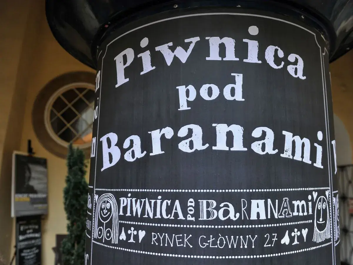 Czarny słup z białym napisem "Piwnica pod Baranami" i ozdobnymi elementami.