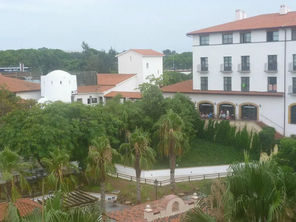 Edificio blanco con tejado naranja, palmeras y vegetación exuberante. Parece el hotel El Paso PortAventura, listo para recibir a sus huéspedes.