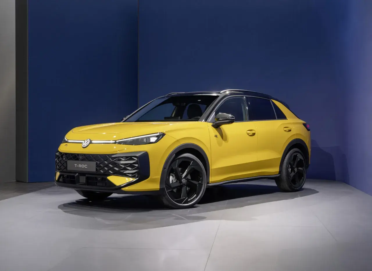 Żółty Volkswagen T-Roc VW z czarnym dachem i felgami stoi na szarej podłodze przed niebieską ścianą.