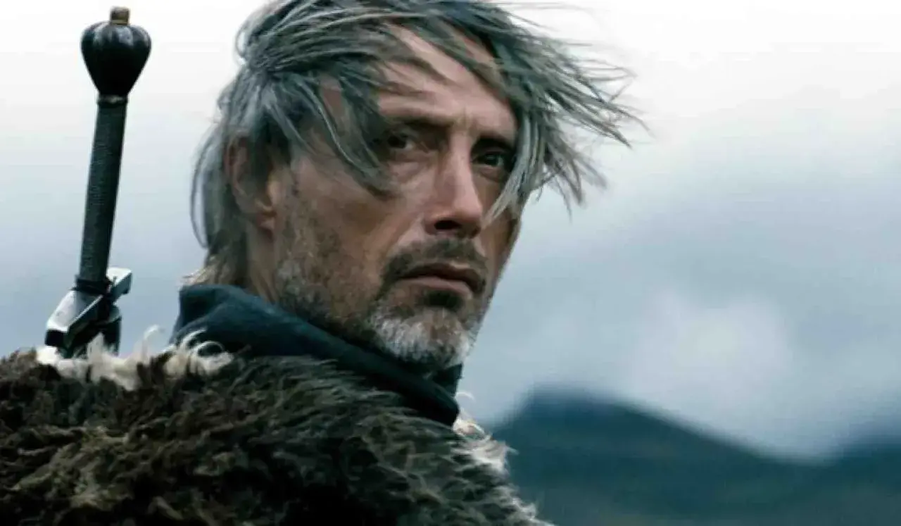 Mads Mikkelsen młody, z mieczem za plecami i w futrzanym płaszczu, spogląda w dal.