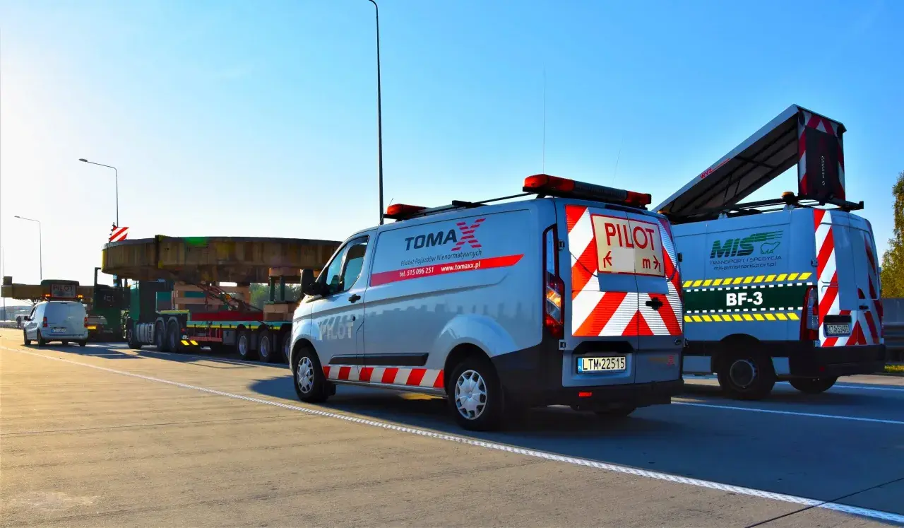 Pojazd ponadgabarytowy transportowany na platformie, eskortowany przez dwa samochody pilotujące z napisem "PILOT".