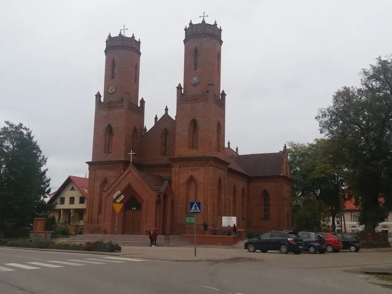 Imponujący kościół w Krokowej z cegły, z dwiema wieżami i zegarami, otoczony zielenią i samochodami.